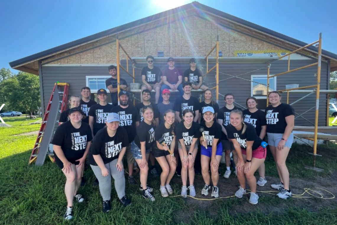 A group of young people in black shirts smiles together in front of a house