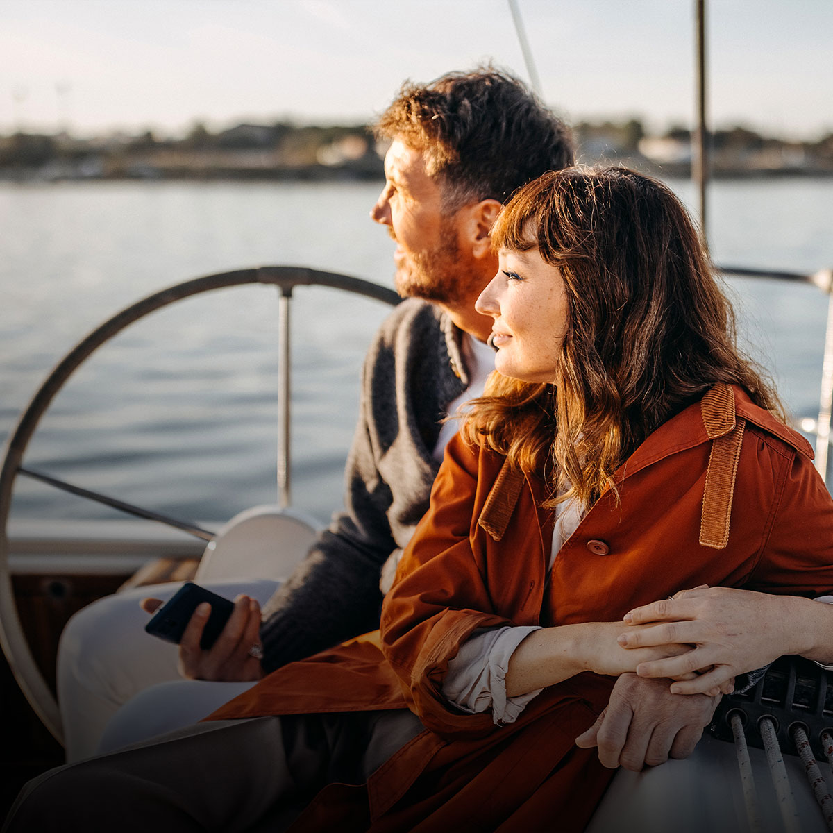 A couple sits on a sailboat, gazing at the serene water during sunset. The woman wears an orange coat, creating a relaxed and peaceful mood.