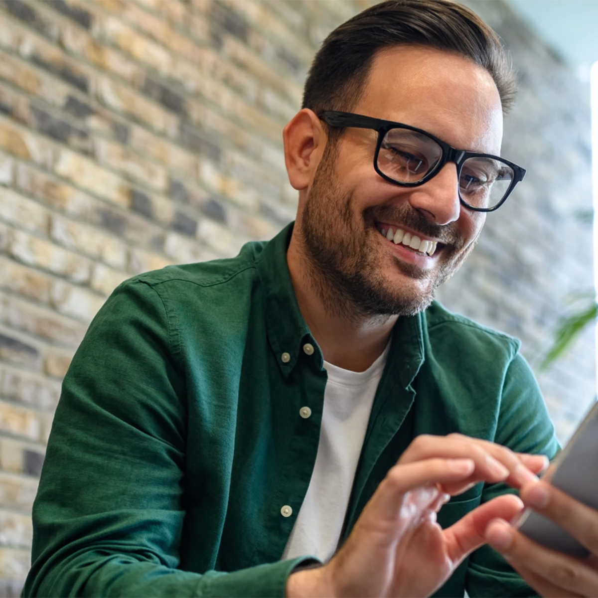  A smiling man in glasses is using his phone, appearing engaged and happy in a casual setting.